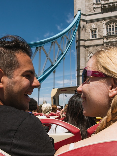 Big Bus London crossing Tower Bridge with Thames River cruise boat visible.