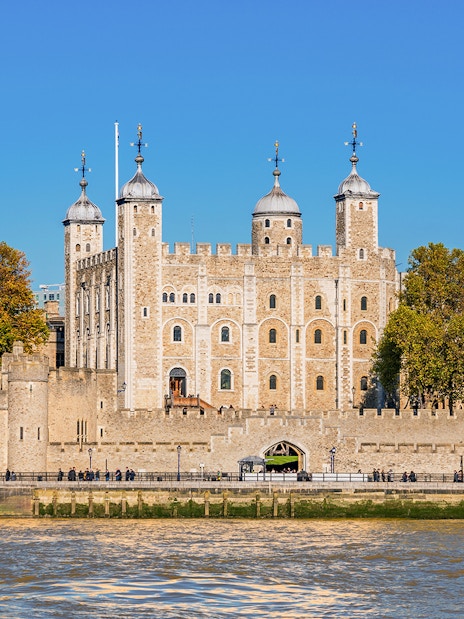 Tower of London exterior with tourists and Thames River boat in view.